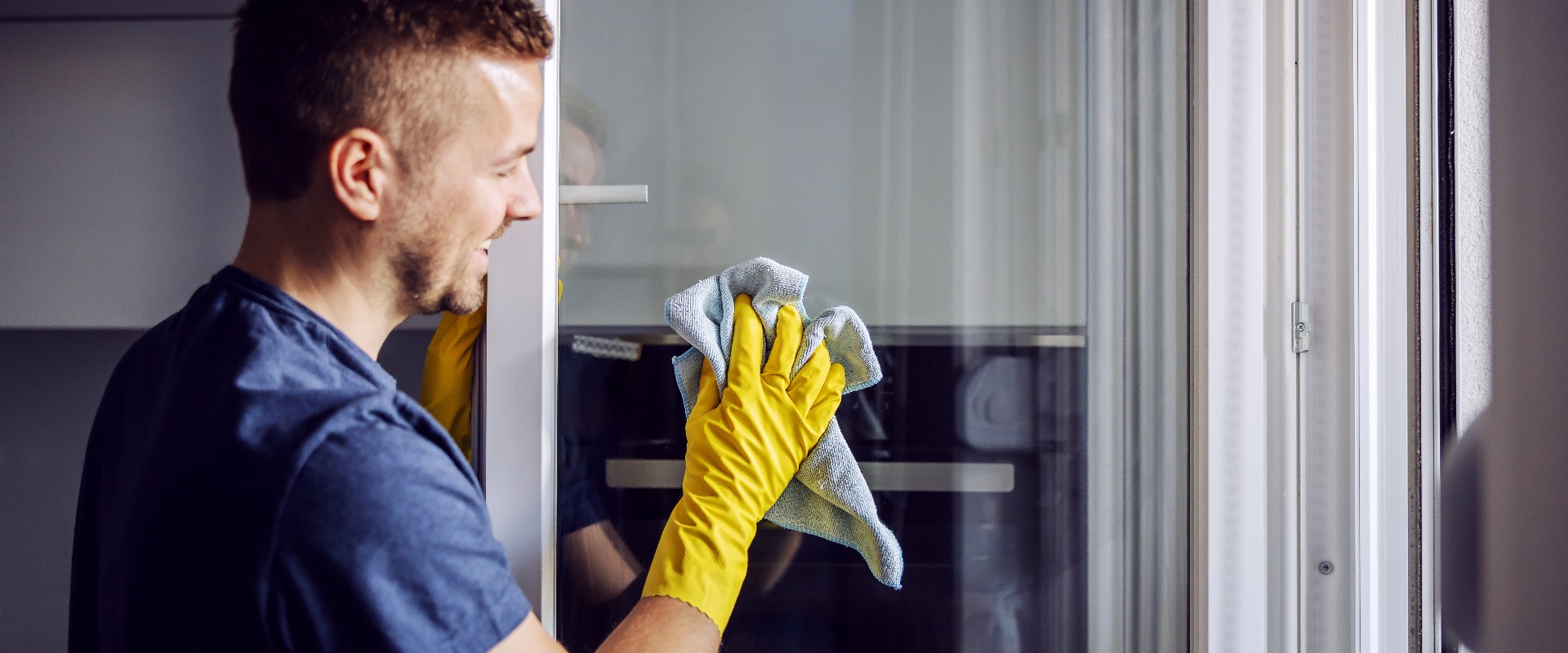 Junger bärtiger lächelnder positiver Mann mit Gummihandschuhen, der ein Fenster mit einem Tuch putzt.
