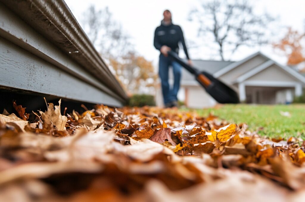 Ein Landschaftsgärtner säubert mit einem Laubbläser den Rand eines Hauses von herabgefallenem Herbstlaub.